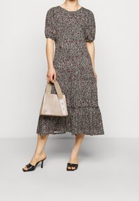 Vestido negro con patrón floral blanco y rojo, con mangas cortas abullonadas y falda de volantes. La modelo sostiene un bolso de mano texturizado en color beige con asas cortas.