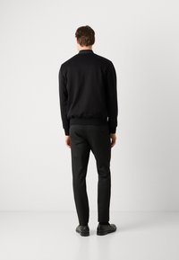 Čierna bomber bunda s rebrovanými manžetami a lemom, nosená s priehľadnými čiernymi nohavicami a elegantnými čiernymi topánkami. Jednoduchá, jednofarebná, s minimálnym detailovaním.