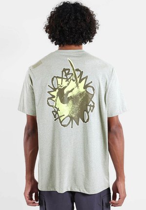 Homme aux cheveux bouclés portant un T-shirt gris clair avec un dessin d'un surfeur chevauchant une vague et un texte circulaire au dos.