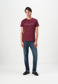 Młody mężczyzna stojący przy białej ścianie, ubrany w bordo T-shirt Tommy Hilfiger, niebieskie dżinsy i czarne mokasyny z frędzlami.