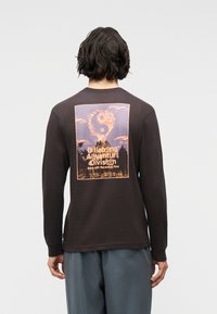 Άτομο με σκούρα μαλλιά φοράει μαύρο μακρυμάνικο πουκάμισο με ένα γραφικό ηφαίστειου, όπου ο καπνός σχηματίζει το σύμβολο γιν-γιανγκ, και κείμενο στην πλάτη.