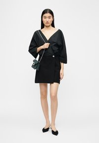 Femme portant un chemisier noir à col en V profond, une mini-jupe noire avec boutons, des chaussures noires à bouts pointus et un petit sac bandoulière vert.