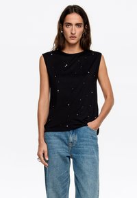 Czarna, bezrękawnikowa bluzka z miękkiego materiału, ozdobiona małymi metalowymi aplikacjami. W zestawie z jasnoniebieskimi, szerokimi spodniami typu jeans.