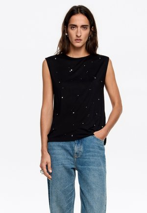 Czarna, bezrękawnikowa bluzka z miękkiego materiału, ozdobiona małymi metalowymi aplikacjami. W zestawie z jasnoniebieskimi, szerokimi spodniami typu jeans.