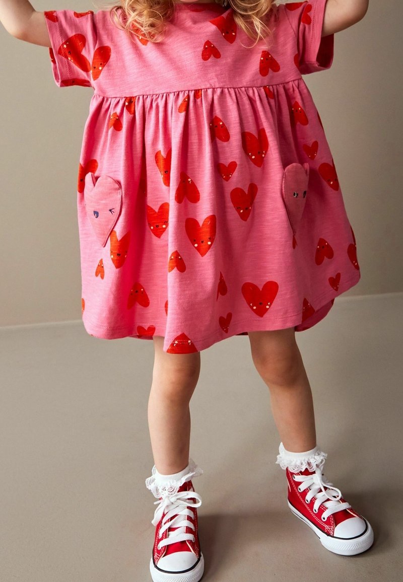 Jeune enfant portant une robe rose avec des motifs de cœurs rouges et des baskets montantes rouges avec des chaussettes blanches à dentelle, debout sur un sol neutre.