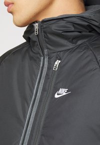 Black nylon jacket with zippered front, high collar, and reflective accents. Features a white Nike logo on the left chest.