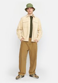 Beige corduroy shirtjack, olijf groene t-shirt, bruine wijde broek en olijf groene bucket hat. Geribbelde materialen met contrasterende kleuren.