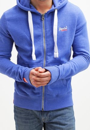 Mann trägt einen blauen Kapuzenpullover mit Reißverschluss und weißen Kordeln sowie Jeans, Hände auf Taillenhöhe verschränkt.