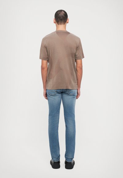 Kortermet brun T-skjorte med en teksturert, mønstret overflate, kombinert med lyseblå jeans og svarte sko. Synlig søm på skjorten.