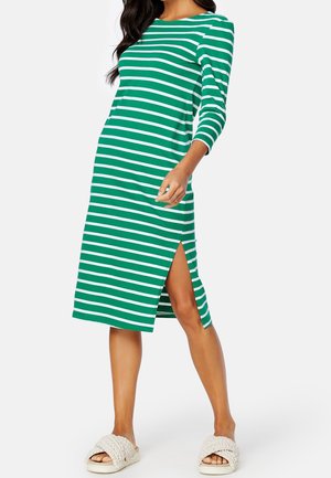 Vrouw draagt een midi-jurk met groene en witte strepen, lange mouwen en een zijsplit, gecombineerd met witte geweven slippers.