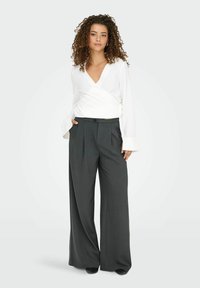 Femme aux cheveux bouclés portant un chemisier blanc croisé et un pantalon gris foncé taille haute à jambes larges, debout avec une main dans la poche.