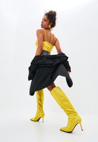Top scurt din satin galben, fustă plisată neagră și cizme galbene cu ținte, până la genunchi, cu fermoare. Modelul pozează cu spatele întors și cu părul aranjat.