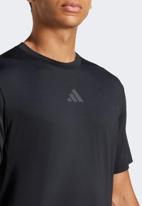 Camiseta deportiva negra hecha de un tejido ligero y suave. Presenta un cuello redondo, mangas cortas y un discreto logo de trébol en el pecho.