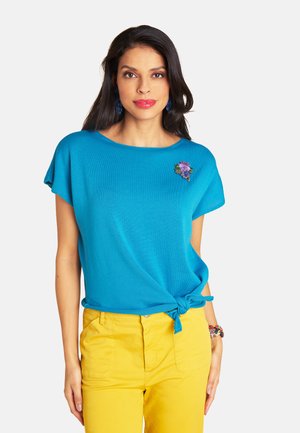 Woman wearing a blue short-sleeve top with knotted hem and floral brooch, paired with yellow pants and colorful bracelet.