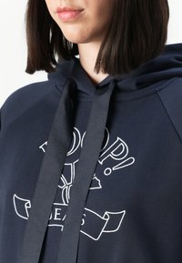 Ženska v mornarsko modrem hoodie-ju s širokimi vrvica in belim vezenim grafičnim dizajnom na prsih.