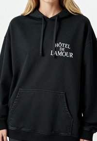 Fekete, oversized kapucnis pulcsi, pamutból készült, elülső zsebbel és húzózsinórral. Fehér hímzett felirattal rendelkezik: "HÔTEL DE L'AMOUR."