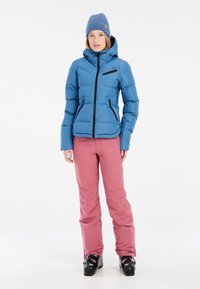 Hellblaues Puffer-Jacket mit schwarzen Reißverschlüssen und Taschen, kombiniert mit rosa Schneehosen und schwarzen Skischuhen; hat eine Kapuze und einen Reißverschluss.