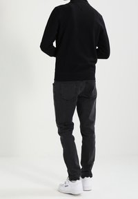 Sudadera negra con cuello alto, vaqueros negros a juego y zapatillas blancas. El material parece suave, con un ajuste relajado y detalles mínimos.