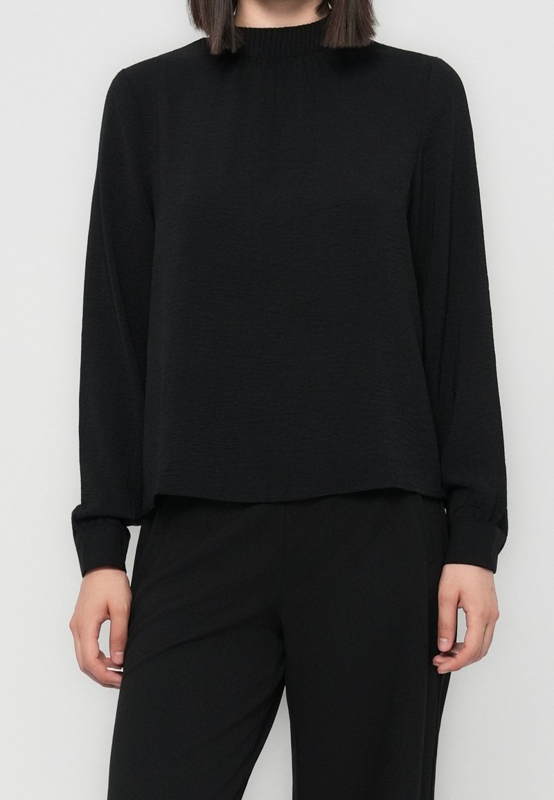Personne portant un haut noir à manches longues avec un col montant et un pantalon noir, debout devant un fond clair uni.