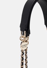 Svart läderväska med guldtonad metallhardware, kedje-accenter och präglad logotag som läser "GUESS EST.1981." Slät textur.
