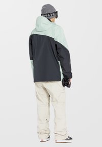 Chaqueta de nieve para hombre en verde menta y negro, con capucha, mangas largas y puños elásticos, combinada con pantalones de nieve beige y zapatillas negras.