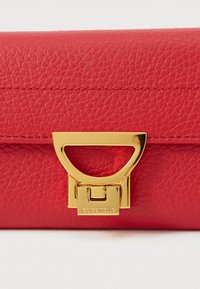 Červená texturovaná kožená peněženka se zlatým kovovým uzávěrem, na kterém je vyryto "COCCINELLE", umístěným na bílém pozadí.