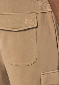 Shorts cargo beiges en tissu texturé, avec une poche cousue portant un logo en relief. Coutures visibles le long des coutures.
