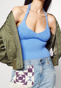 Žena v modrém pleteném topu bez rukávů, olivově zelené bundě přehozené přes ramena a světle modrých džínách se vzorovanou látkovou záplatou.