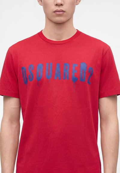 Jeune personne portant un t-shirt rouge avec des lettres bleues dégoulinantes formant le mot DSQUARED2 sur la poitrine.