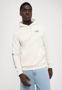 Man wearing a cream EA7 Emporio Armani hoodie with front pocket and logo on chest and sleeve paired with light blue jeans.