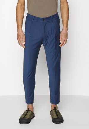 Uomo con pantaloni blu navy aderenti e scarpe slip-on verde oliva in piedi contro uno sfondo chiaro uniforme.