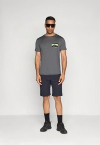 Harmaa t-paita, jossa on rintatasku ja vihreä-musta kuvio, yhdistettynä tummansinisiin shortseihin, mustiin sukkiin ja tummiin jalkineisiin.