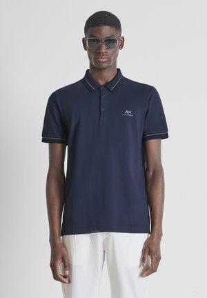 Jonge man in een donkerblauw poloshirt en witte broek, staand tegen een effen lichte achtergrond, met een zonnebril op zijn gezicht.