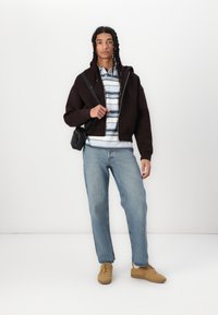 Haut à capuche zippé marron sur une chemise rayée, jeans bleu clair et baskets beige. Comprend un petit sac bandoulière noir.