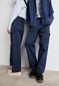 Donkerblauwe wijde broek met zichtbare zakdetails en stiksels; gecombineerd met een lichtblauw overhemd met knopen en een denimjack.