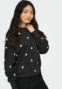 Sieviete ar gariem tumšiem matiem, ģērbusies melnā sportiskā džemperī ar maziem baltiem margrietiņu rakstiem, pozē ar vienu roku pie sava kakla.