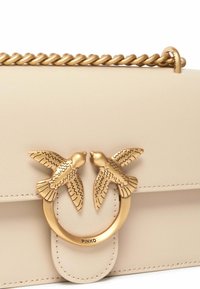 Borsa in pelle beige con tracolla a catena dorata, un chiusura circolare con due dettagli a forma di uccello e bordi cuciti.