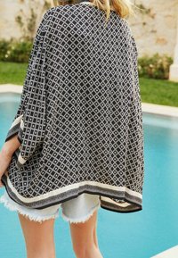 Sort og hvid mønstret poncho med diamantdesign, der har en tekstureret kant og kontrastkant, båret over lyse denimshorts.