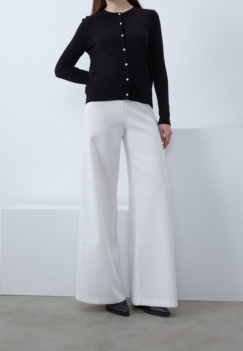 Femme portant un cardigan à manches longues noir boutonné et un pantalon blanc taille haute à jambes larges, debout sur un sol gris contre un mur blanc.