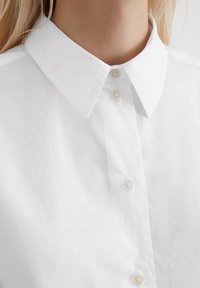Camicia bianca in cotone con colletto classico, dotata di una chiusura a bottoni e piccoli bottoni arrotondati, che mostra una texture liscia e un design pulito.