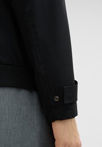 Schwarze Bomberjacke mit geripptem Bündchen, ausgestattet mit einem Druckknopfdetail und subtilen Glanz, präsentiert vor einem grauen strukturierten Stoffhintergrund.