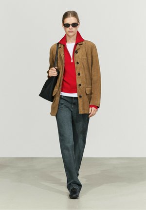 Donna che cammina indossando occhiali da sole neri, giacca in pelle scamosciata marrone, maglione rosso con zip, jeans scuri, scarpe nere e porta una borsa a tracolla nera.