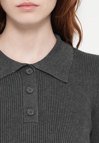 Mørkegrå ribbestrikket polo genser med krage og tre matchende knapper foran. Den strukturerte teksten fremhever designet.