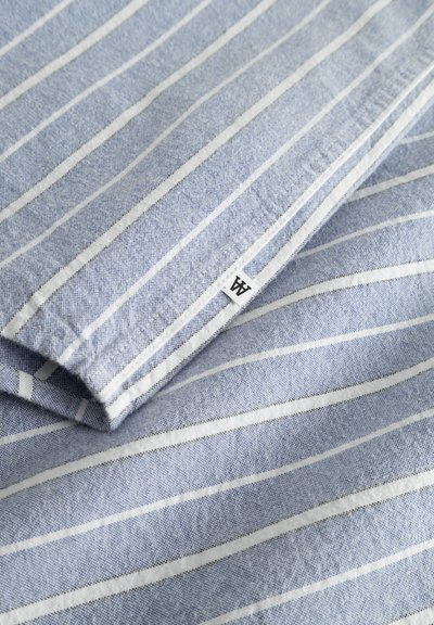 Tissu bleu clair avec des rayures blanches, plié. Comprend une petite étiquette avec un logo. La texture semble douce et lisse.