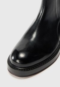 Bota de tornozelo em couro preto com acabamento brilhante, ponta arredondada e sola robusta e texturizada. Apresenta costura contrastante ao longo da borda.