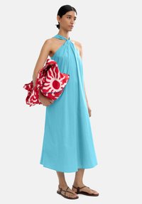 Sukienka w kolorze jasnego turkusu z dekoltem halter, wykonana z lekkiego materiału. Modelka trzyma czerwoną torebkę z motywem słońca.
