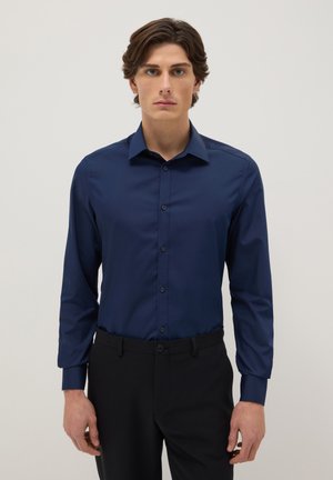 Jeune homme aux cheveux bruns portant une chemise ajustée bleu marine à boutons et un pantalon noir, sur un fond clair uni.