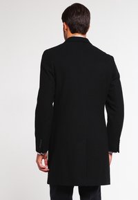 Man in een lange zwarte jas met dichtgeknoopte mouwen, staand met de rug naar de camera tegen een effen witte achtergrond.