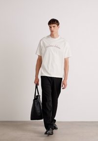 White cotton t-shirt with "calvin klein" logo, paired with black trousers and carrying a black tote bag, walking on a neutral background.