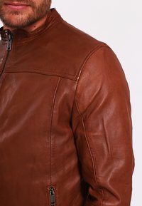 Giacca in pelle marrone con colletto alto, design aderente e tasche laterali con zip. Texture liscia con dettagli di cucitura minimi.